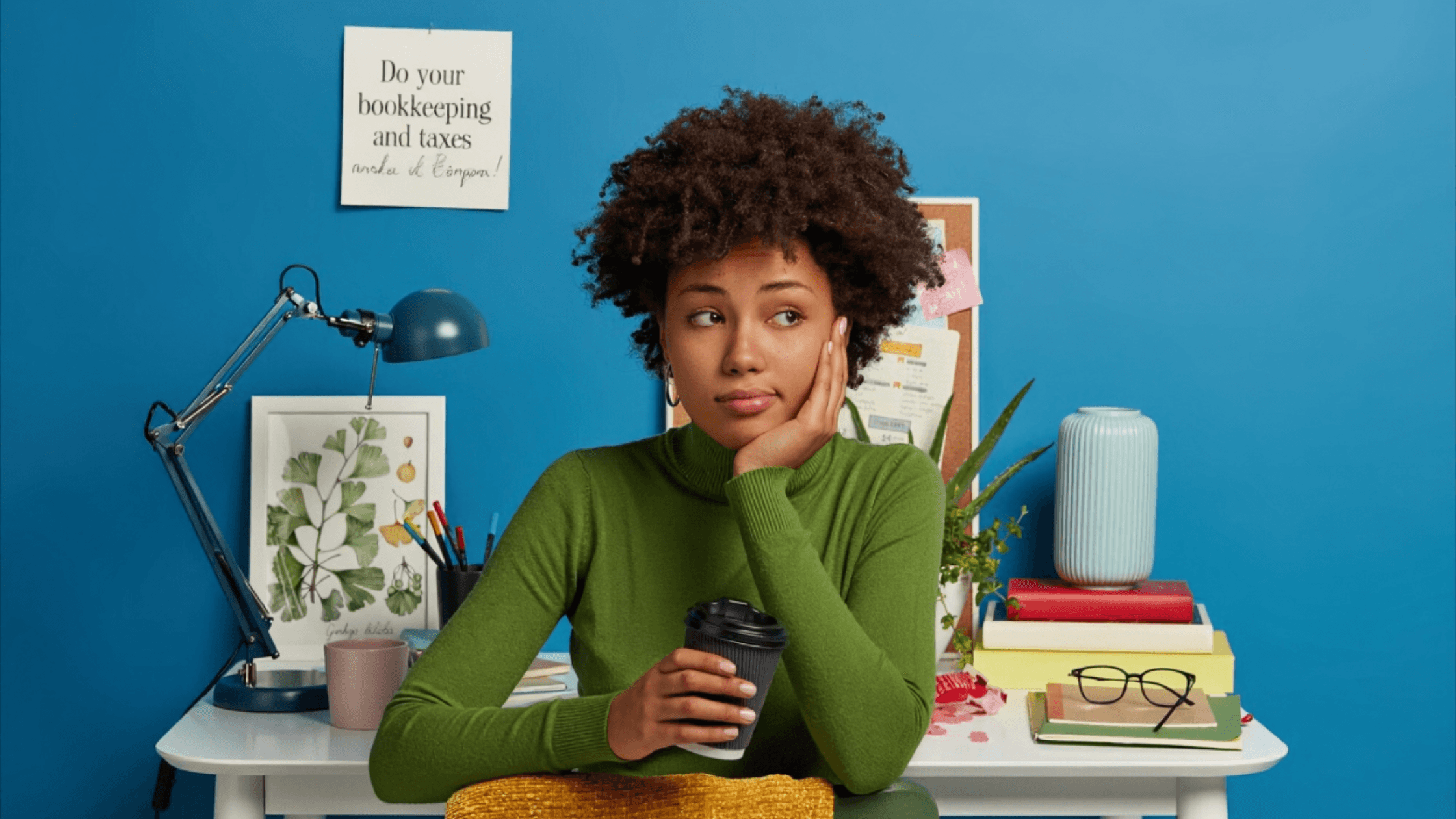 A young creator sits at a desk holding a coffee cup and looking worried about bookkeeping and tax