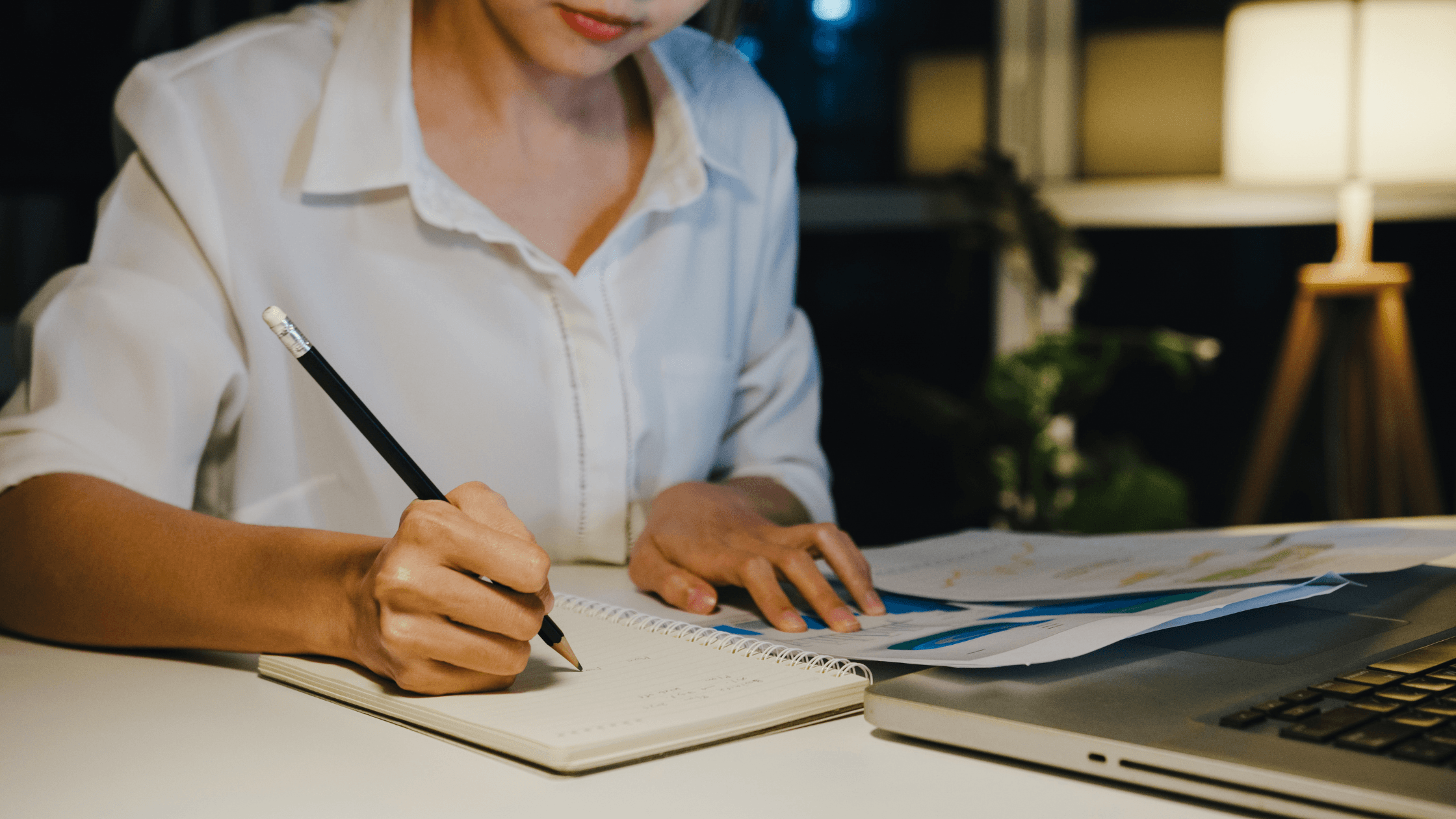 Person writing estimated tax calculations in a notebook beside a laptop and financial documents at a desk.
